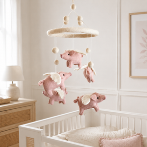 Felt flying pig baby mobile in pink and cream hanging above a light wood crib in a sunlit nursery.