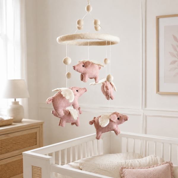 Felt flying pig baby mobile in pink and cream hanging above a light wood crib in a sunlit nursery.