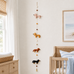 Felt dachshund garland with four colored dogs and pom bead accents hanging on a soft white nursery wall.