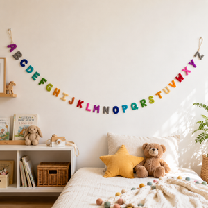 Rainbow felt alphabet garland strung above a child's bed with a teddy bear and star pillow nearby
