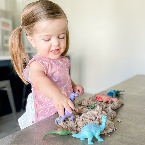 Blush Dino Friends Bapron for babies, angled view showcasing playful dinosaur design and practical bib features.