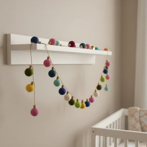 Felt color ball lightbulb garland draped along a white nursery shelf in soft daylight