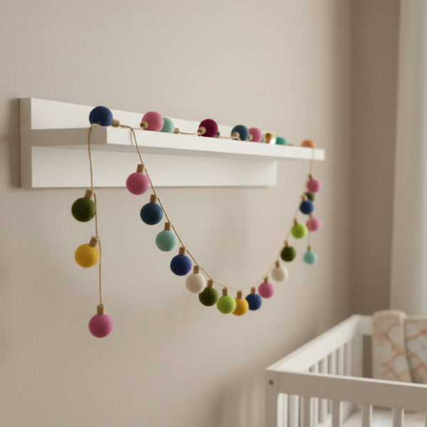 Felt color ball lightbulb garland draped along a white nursery shelf in soft daylight