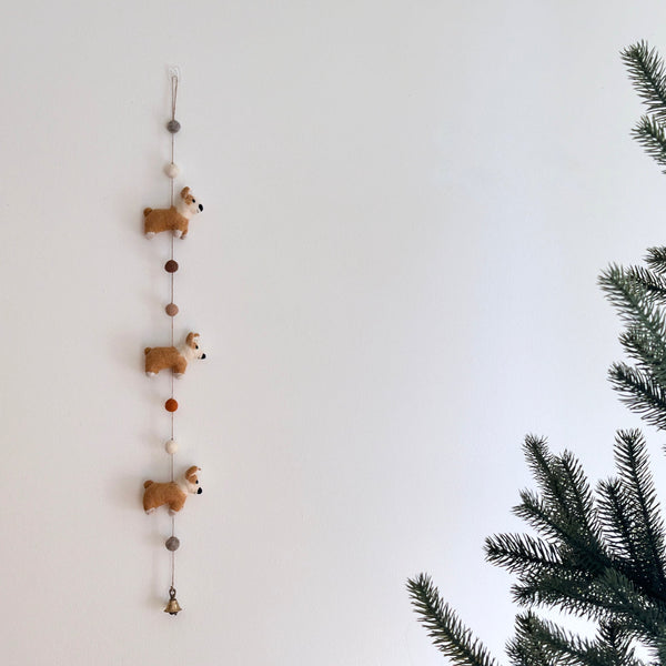 Felt corgi garland hanging on a white wall near pine branches, full length view
