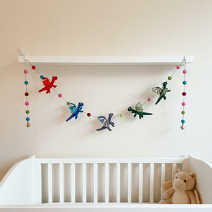Felt dragon garland draped along a nursery shelf above a crib in soft daylight