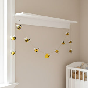 Felt bee garland draped along a white nursery shelf, soft daylight and neutral walls in the room