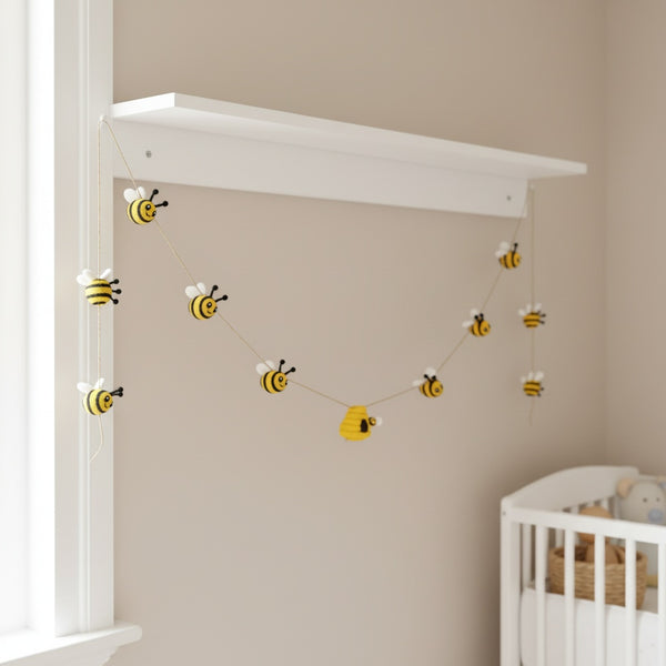 Felt bee garland draped along a white nursery shelf, soft daylight and neutral walls in the room