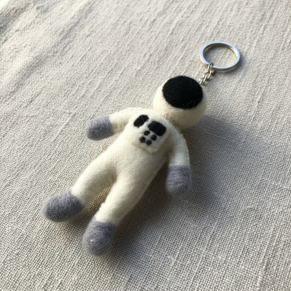 Felt astronaut keychain resting on a linen surface, close-up showing the wool felt texture.