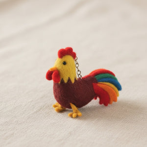 Felt rooster keychain resting on light linen fabric, soft light highlighting its wool texture.