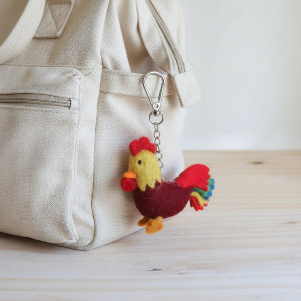 Felt rooster keychain clipped to a canvas diaper bag on a wooden table in soft daylight.