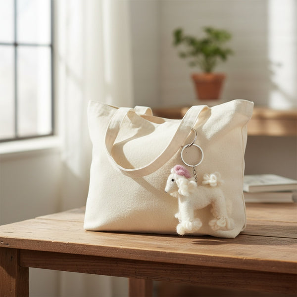 White felt poodle keychain clipped to a canvas diaper bag on a wooden table in soft daylight.