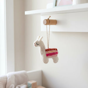 Felt llama ornament hanging from a wooden peg on a white nursery shelf in soft daylight