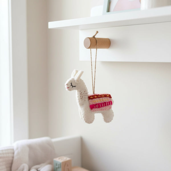 Felt llama ornament hanging from a wooden peg on a white nursery shelf in soft daylight