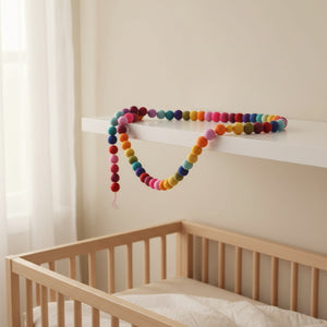 Felt rainbow pompom garland draped along a white nursery shelf in soft daylight.