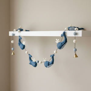 Felt whale garland draped along a white nursery shelf, soft daylight on the blue wool whales