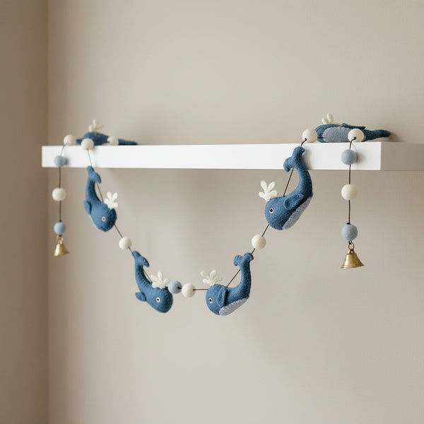Felt whale garland draped along a white nursery shelf, soft daylight on the blue wool whales